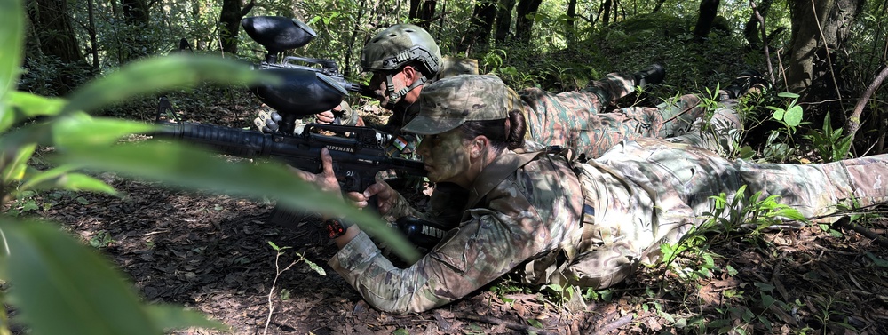 Multiple individuals in camouflage military-style uniforms are lying prone in a forested environment, aiming Tippmann-brand paintball markers forward. The scene appears to be a military training exercise or tactical paintball/airsoft simulation in a dense woodland setting. The participants are in a line formation, suggesting coordinated tactical training.