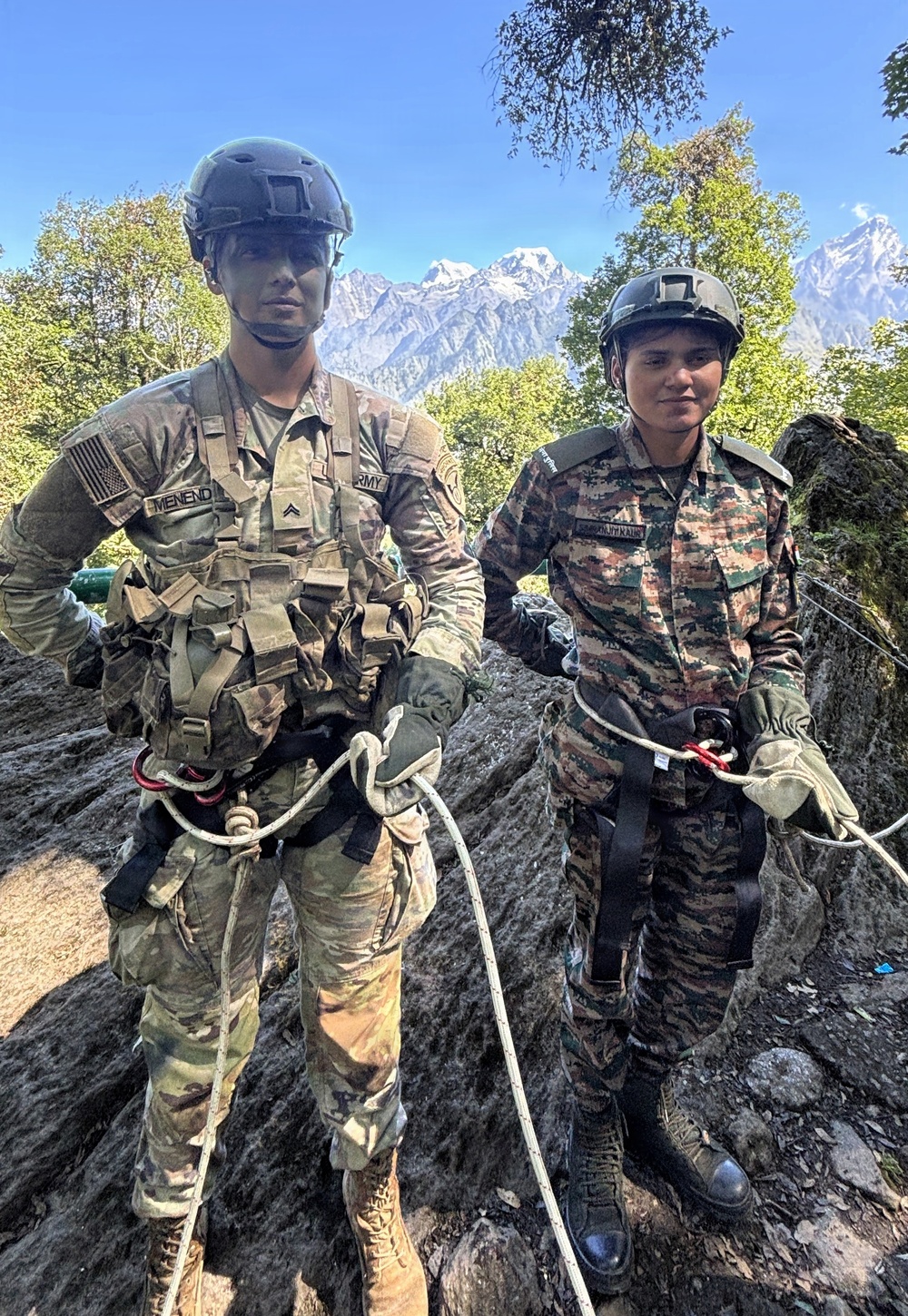 Two soldiers in military uniforms are standing on rocky terrain with rappelling/climbing harnesses and ropes attached. One soldier wears US Army MultiCam (OCP) uniform with an American flag patch and name tape reading 'MENEND', while the second wears a different camouflage pattern consistent with Indian Army or similar South Asian military uniform. Snow-capped mountains are visible in the background, suggesting a high-altitude joint military training exercise.