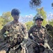Two soldiers in military uniforms are standing on rocky terrain with rappelling/climbing harnesses and ropes attached. One soldier wears US Army MultiCam (OCP) uniform with an American flag patch and name tape reading 'MENEND', while the second wears a different camouflage pattern consistent with Indian Army or similar South Asian military uniform. Snow-capped mountains are visible in the background, suggesting a high-altitude joint military training exercise.