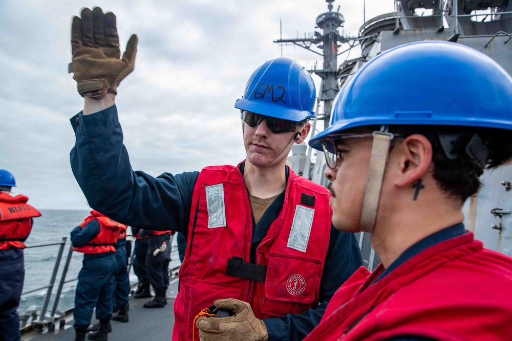 Two naval personnel wearing blue hard hats and red survival/life vests are on the deck of a military vessel at sea. The sailor in the center has 'GM2' written on his hard hat and is raising a gloved hand, appearing to give a signal or instruction. Additional crew members in similar safety gear are visible in the background.