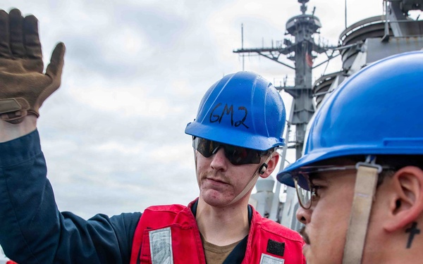 Two naval personnel wearing blue hard hats and red survival/life vests are on the deck of a military vessel at sea. The sailor in the center has 'GM2' written on his hard hat and is raising a gloved hand, appearing to give a signal or instruction. Additional crew members in similar safety gear are visible in the background.
