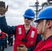 Two naval personnel wearing blue hard hats and red survival/life vests are on the deck of a military vessel at sea. The sailor in the center has 'GM2' written on his hard hat and is raising a gloved hand, appearing to give a signal or instruction. Additional crew members in similar safety gear are visible in the background.