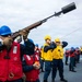 A naval crew member wearing a red life vest and blue hard hat is firing a line-throwing rifle from the deck of a warship, launching a visible red line across the water. Multiple other crew members in yellow and blue hard hats with life vests stand behind, covering their ears during the shot. This appears to be a replenishment-at-sea (RAS) or ship-to-ship line transfer exercise or operation.