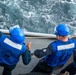 Two naval personnel wearing blue safety vests and blue hard hats are working on the deck of a ship at sea. They appear to be handling lines or ropes during what looks like an underway replenishment or line-handling evolution. The churning wake of the vessel is visible in the background, indicating the ship is underway.