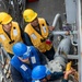 Four personnel wearing safety equipment are working on large pipe fittings and valves on what appears to be the deck of a naval or industrial vessel. Two individuals wear yellow hard hats and yellow life vests, while two others wear blue hard hats and blue coveralls. They appear to be conducting a replenishment-at-sea (RAS) or maintenance operation involving a large flanged pipe coupling.