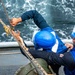 Three personnel wearing blue hard hats and blue uniforms are working together on the deck of a naval vessel, handling large mooring or rigging ropes. The workers appear to be engaged in a line-handling evolution, likely during an underway replenishment or mooring operation. Churning ocean water is visible in the background, indicating the vessel is underway.