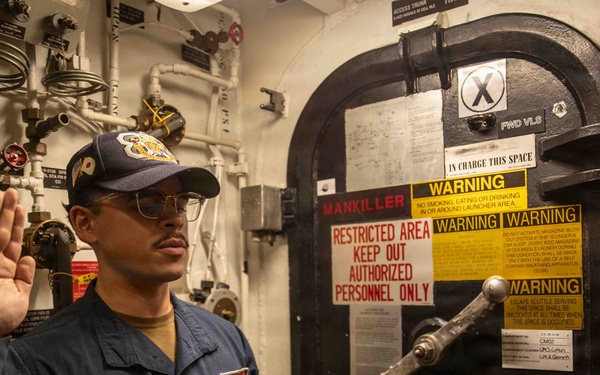 A U.S. Navy sailor in blue coveralls is raising his right hand, appearing to take an oath or re-enlistment ceremony aboard a naval vessel. The ceremony is taking place in front of a restricted access watertight door marked with multiple warning signs. An officer in khaki uniform faces the sailor, also with a raised hand.