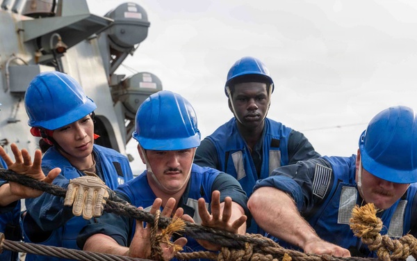 Five naval personnel in blue uniforms and blue hard hats are collectively hauling a large rope on the deck of a military vessel at sea. The crew members appear to be performing a line-handling evolution, likely during an underway replenishment or mooring operation. The overcast sky and open ocean are visible in the background, along with naval ship superstructure.
