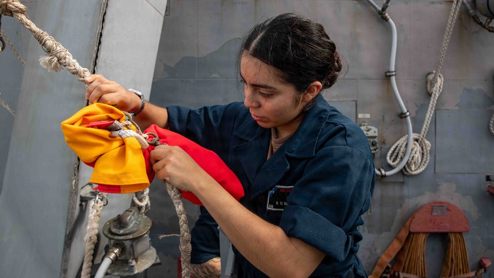 A female sailor in dark blue Navy coveralls is working with signal flags (orange and red) attached to a rope on the deck of a naval vessel. She appears to be securing or preparing nautical signal flags, which are used for maritime communication. The setting is clearly aboard a military ship, with gray metal bulkheads and deck equipment visible in the background.