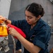 A female sailor in dark blue Navy coveralls is working with signal flags (orange and red) attached to a rope on the deck of a naval vessel. She appears to be securing or preparing nautical signal flags, which are used for maritime communication. The setting is clearly aboard a military ship, with gray metal bulkheads and deck equipment visible in the background.