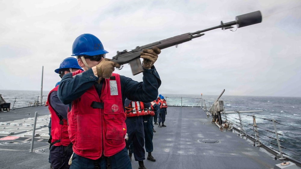 A naval sailor aboard a warship is firing a line-throwing gun (likely an M14 rifle with a line-throwing attachment) on the ship's deck. Several other crew members wearing blue hard hats and red life vests are present on deck observing or assisting. The activity appears to be a replenishment-at-sea (RAS) or line-throwing exercise conducted on open ocean.