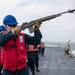 A naval sailor aboard a warship is firing a line-throwing gun (likely an M14 rifle with a line-throwing attachment) on the ship's deck. Several other crew members wearing blue hard hats and red life vests are present on deck observing or assisting. The activity appears to be a replenishment-at-sea (RAS) or line-throwing exercise conducted on open ocean.