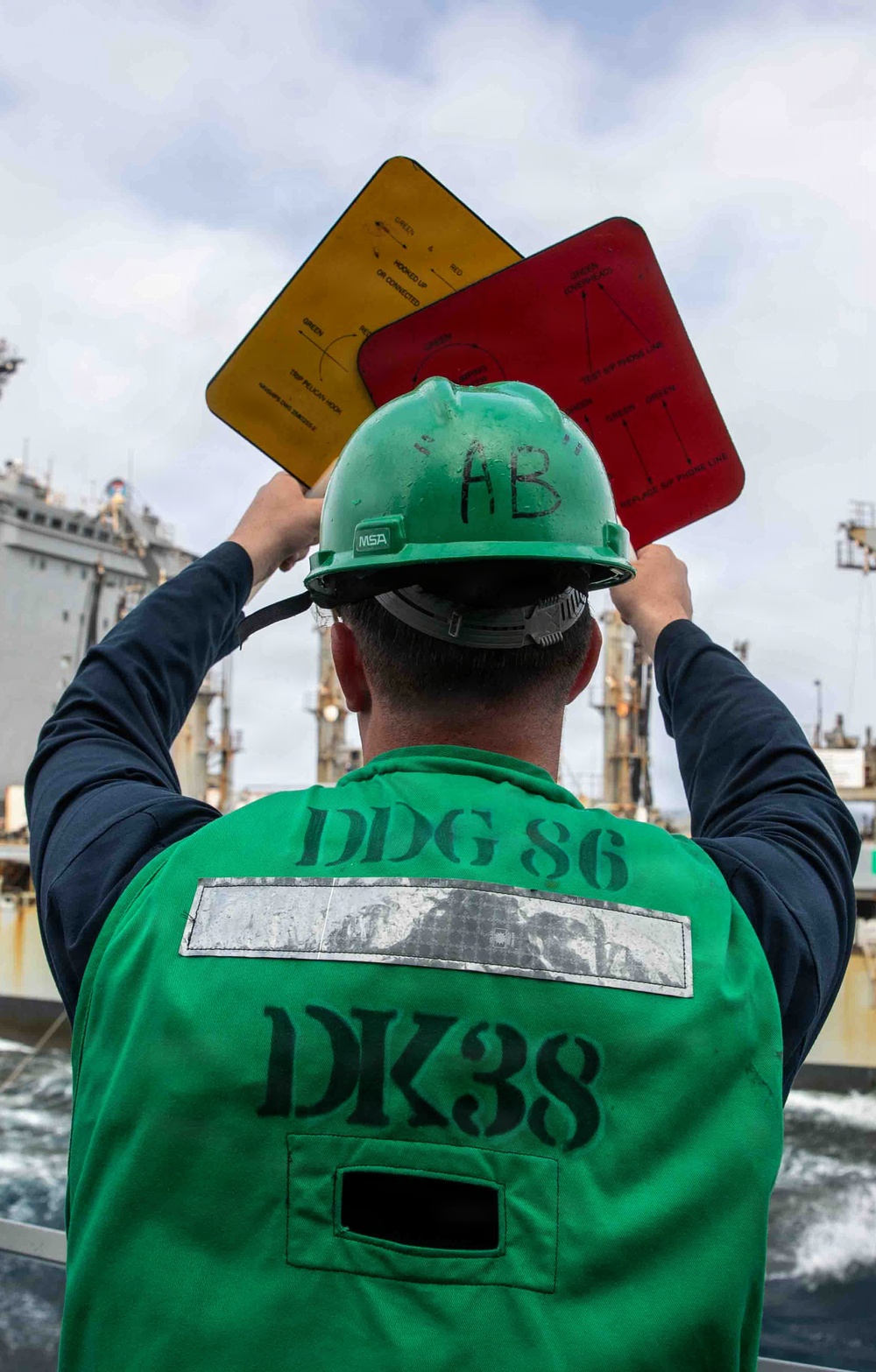 A U.S. Navy sailor assigned to DDG 86 (USS Shoup, an Arleigh Burke-class destroyer) is holding up yellow and red signal boards during an underway replenishment (UNREP) operation at sea. The sailor is wearing a green safety vest marked 'DDG 86' and 'DK38', along with a green MSA hard hat marked 'AB'. A replenishment vessel is visible in the background.