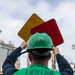 A U.S. Navy sailor assigned to DDG 86 (USS Shoup, an Arleigh Burke-class destroyer) is holding up yellow and red signal boards during an underway replenishment (UNREP) operation at sea. The sailor is wearing a green safety vest marked 'DDG 86' and 'DK38', along with a green MSA hard hat marked 'AB'. A replenishment vessel is visible in the background.
