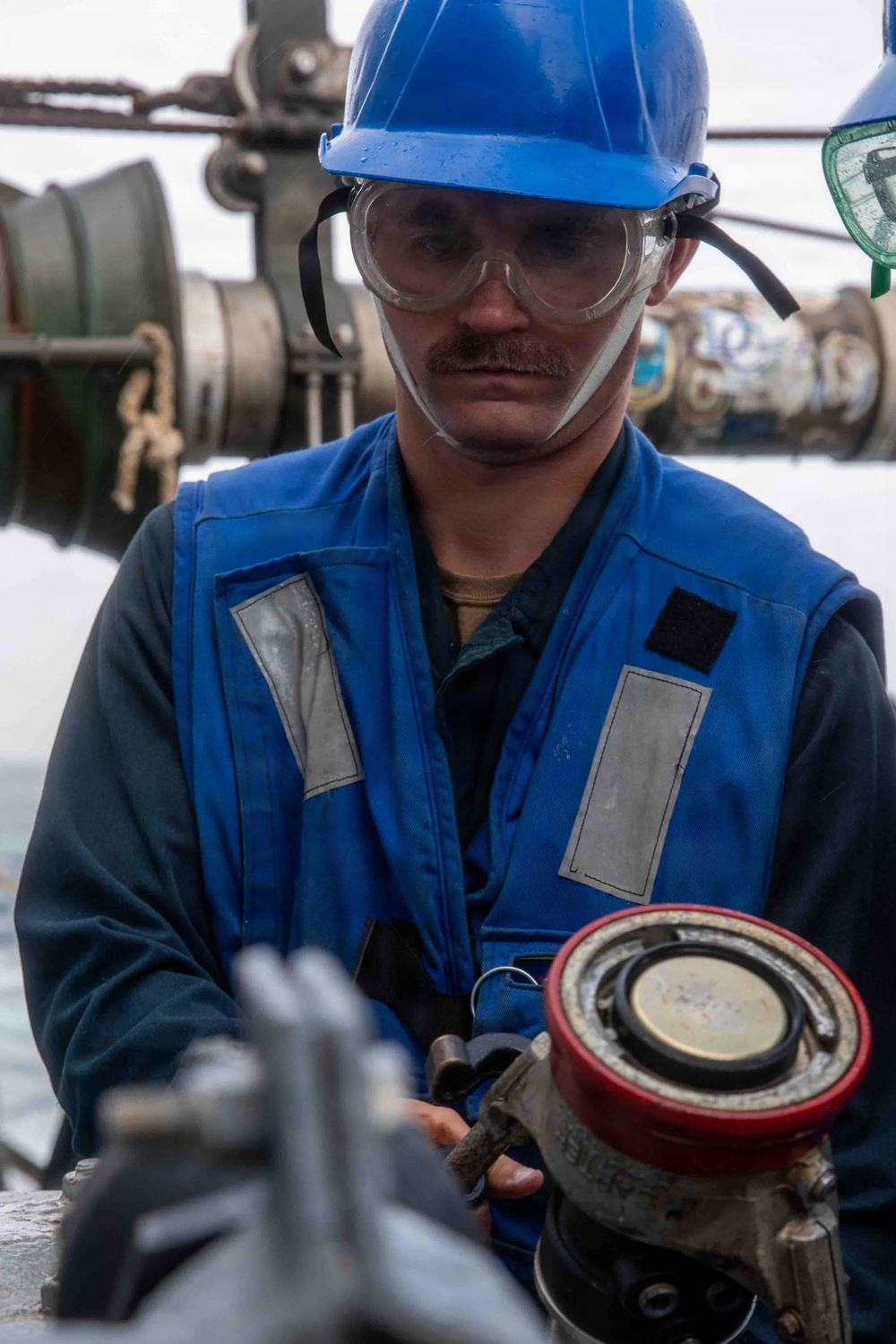 A male worker wearing a blue hard hat, safety goggles, and a blue reflective safety vest is handling industrial equipment, likely a fuel or fluid coupling/nozzle with a red collar. He appears to be on the deck of a ship or offshore platform, surrounded by pipes and mechanical equipment. The worker is focused on the task at hand, suggesting an active maintenance or fueling operation.