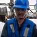 A male worker wearing a blue hard hat, safety goggles, and a blue reflective safety vest is handling industrial equipment, likely a fuel or fluid coupling/nozzle with a red collar. He appears to be on the deck of a ship or offshore platform, surrounded by pipes and mechanical equipment. The worker is focused on the task at hand, suggesting an active maintenance or fueling operation.