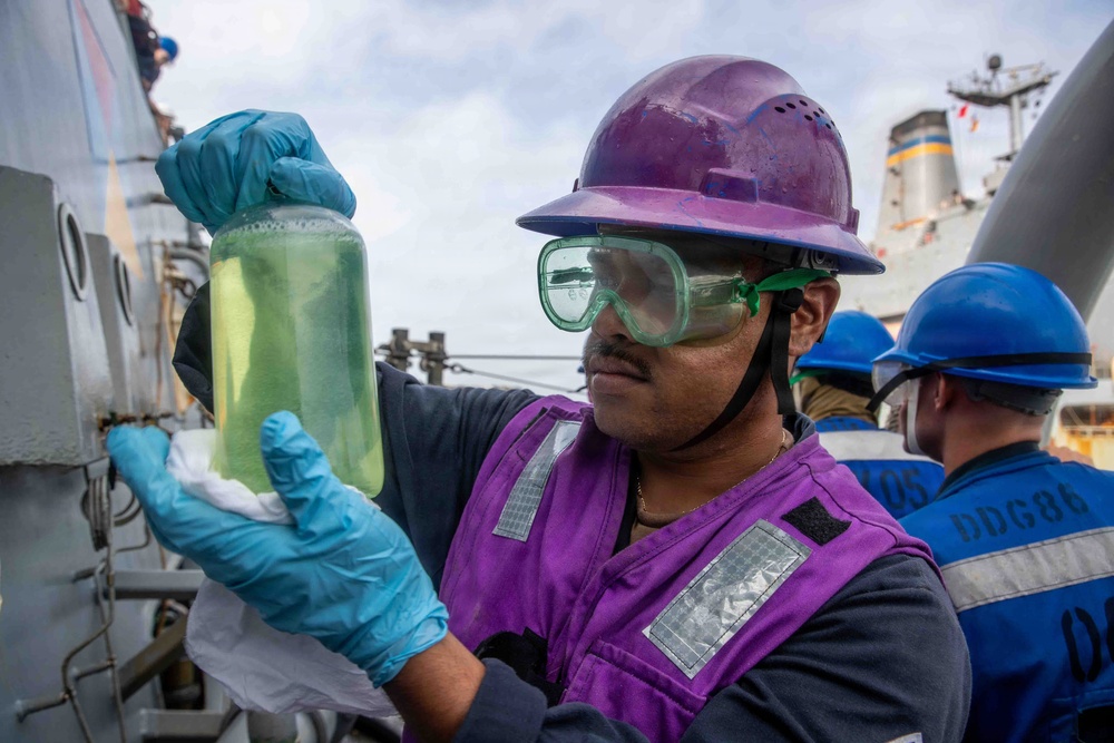 A naval sailor wearing a purple hard hat, safety goggles, purple vest, and blue nitrile gloves is examining a clear jar containing a yellowish-green liquid, likely conducting a fuel or fluid quality inspection. The setting appears to be the deck of a U.S. Navy destroyer, with another crew member visible in the background wearing a blue hard hat. Partial hull markings 'DDG86' are visible on the background sailor's vest, identifying the vessel as USS Shoup or a similar Arleigh Burke-class destroyer.