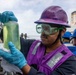 A naval sailor wearing a purple hard hat, safety goggles, purple vest, and blue nitrile gloves is examining a clear jar containing a yellowish-green liquid, likely conducting a fuel or fluid quality inspection. The setting appears to be the deck of a U.S. Navy destroyer, with another crew member visible in the background wearing a blue hard hat. Partial hull markings 'DDG86' are visible on the background sailor's vest, identifying the vessel as USS Shoup or a similar Arleigh Burke-class destroyer.