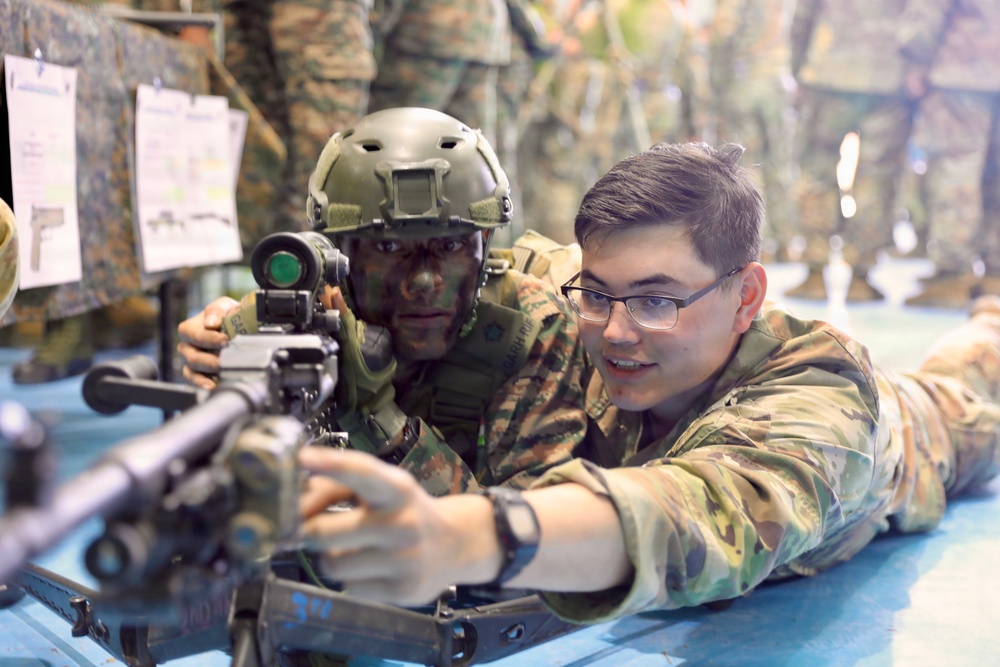 Two military personnel in camouflage uniforms are positioned prone at what appears to be a shooting range or training facility. One soldier wearing a tactical helmet with face paint and a red-dot optic sight is aiming a rifle, while a second soldier wearing glasses appears to be providing instruction or guidance. Additional uniformed personnel are visible standing in the background.