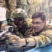 Two military personnel in camouflage uniforms are positioned prone at what appears to be a shooting range or training facility. One soldier wearing a tactical helmet with face paint and a red-dot optic sight is aiming a rifle, while a second soldier wearing glasses appears to be providing instruction or guidance. Additional uniformed personnel are visible standing in the background.