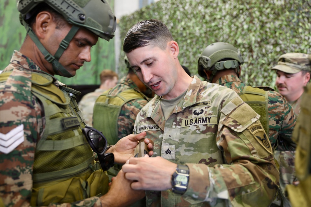 A U.S. Army Sergeant named Rivera-Torres is interacting with a foreign military soldier wearing a different camouflage pattern uniform, appearing to demonstrate or inspect equipment. The setting appears to be a military training or exchange event, indicated by the camouflage netting backdrop and presence of multiple military personnel. The interaction suggests a multinational military cooperation or joint training exercise.