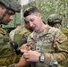A U.S. Army Sergeant named Rivera-Torres is interacting with a foreign military soldier wearing a different camouflage pattern uniform, appearing to demonstrate or inspect equipment. The setting appears to be a military training or exchange event, indicated by the camouflage netting backdrop and presence of multiple military personnel. The interaction suggests a multinational military cooperation or joint training exercise.