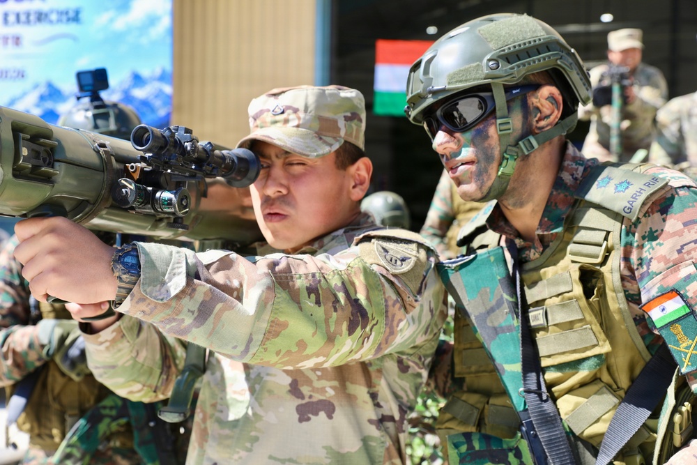 The image depicts a joint military exercise involving soldiers from at least two nations, identifiable by an Indian flag patch and 'GARH RIF' (Garhwal Rifles) unit designation on one soldier's uniform. A soldier in MultiCam/OCP pattern camouflage is aiming a rifle equipped with an optical scope, while an Indian Army soldier in DPCU camouflage with tactical gear and face paint observes closely. A banner and Indian national flag are visible in the background, suggesting a formal bilateral training event.