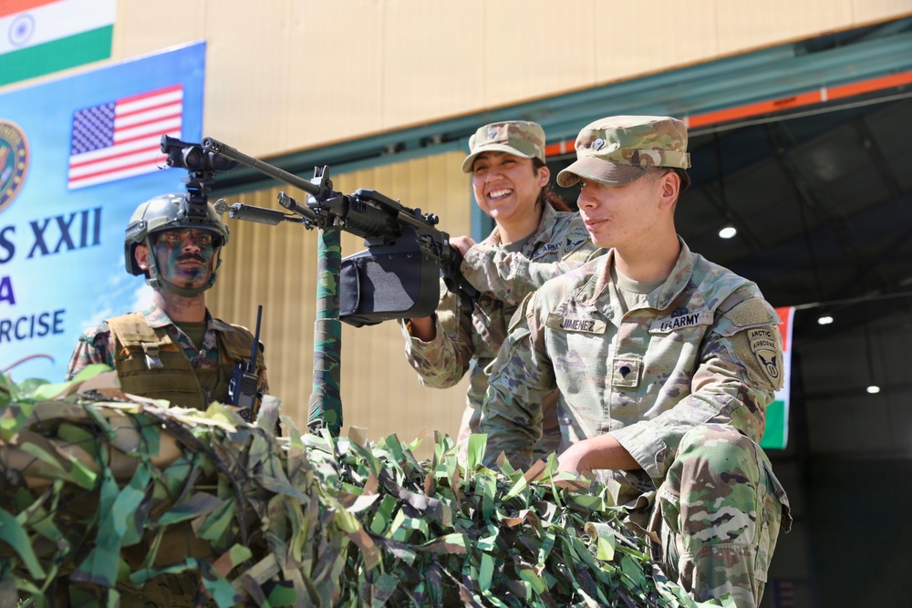 The image shows a joint military exercise between U.S. Army and Indian Army personnel, as indicated by the flags and banner visible in the background reading 'S XXII' and 'EXERCISE'. Two U.S. Army soldiers in OCP (Operational Camouflage Pattern) uniforms are positioned behind a camouflage-netted emplacement with a mounted machine gun, alongside an Indian soldier in combat face paint and helmet. The setting appears to be a demonstration or training event.
