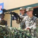The image shows a joint military exercise between U.S. Army and Indian Army personnel, as indicated by the flags and banner visible in the background reading 'S XXII' and 'EXERCISE'. Two U.S. Army soldiers in OCP (Operational Camouflage Pattern) uniforms are positioned behind a camouflage-netted emplacement with a mounted machine gun, alongside an Indian soldier in combat face paint and helmet. The setting appears to be a demonstration or training event.