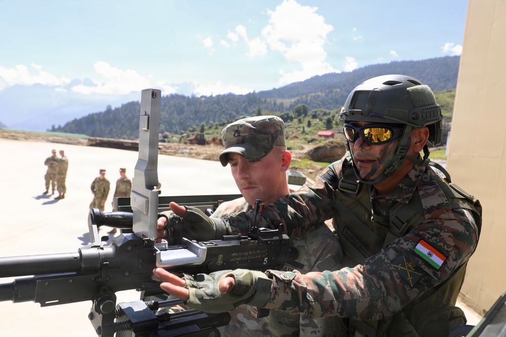 Two soldiers, one wearing an Indian Army uniform with a tricolor flag patch and tactical helmet, and another in what appears to be US Army camouflage, are jointly examining or operating a mounted machine gun. The setting appears to be an outdoor military training area with mountainous terrain in the background. Additional soldiers in US Army uniforms are visible in the background, suggesting a joint military exercise or training event.