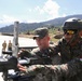 Two soldiers, one wearing an Indian Army uniform with a tricolor flag patch and tactical helmet, and another in what appears to be US Army camouflage, are jointly examining or operating a mounted machine gun. The setting appears to be an outdoor military training area with mountainous terrain in the background. Additional soldiers in US Army uniforms are visible in the background, suggesting a joint military exercise or training event.