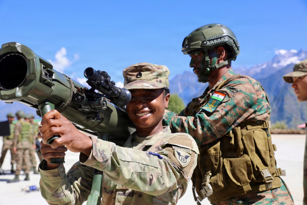 The image shows military personnel from what appears to be a joint training exercise between U.S. Army and Indian Army soldiers. A smiling U.S. soldier in MultiCam uniform is handling a shoulder-fired rocket launcher (appears to be an AT4 or similar weapon system), while an Indian Army soldier in distinctive camouflage with an Indian flag patch assists. The setting appears to be a mountainous region, likely in northern India based on the terrain and context.