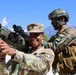 The image shows military personnel from what appears to be a joint training exercise between U.S. Army and Indian Army soldiers. A smiling U.S. soldier in MultiCam uniform is handling a shoulder-fired rocket launcher (appears to be an AT4 or similar weapon system), while an Indian Army soldier in distinctive camouflage with an Indian flag patch assists. The setting appears to be a mountainous region, likely in northern India based on the terrain and context.