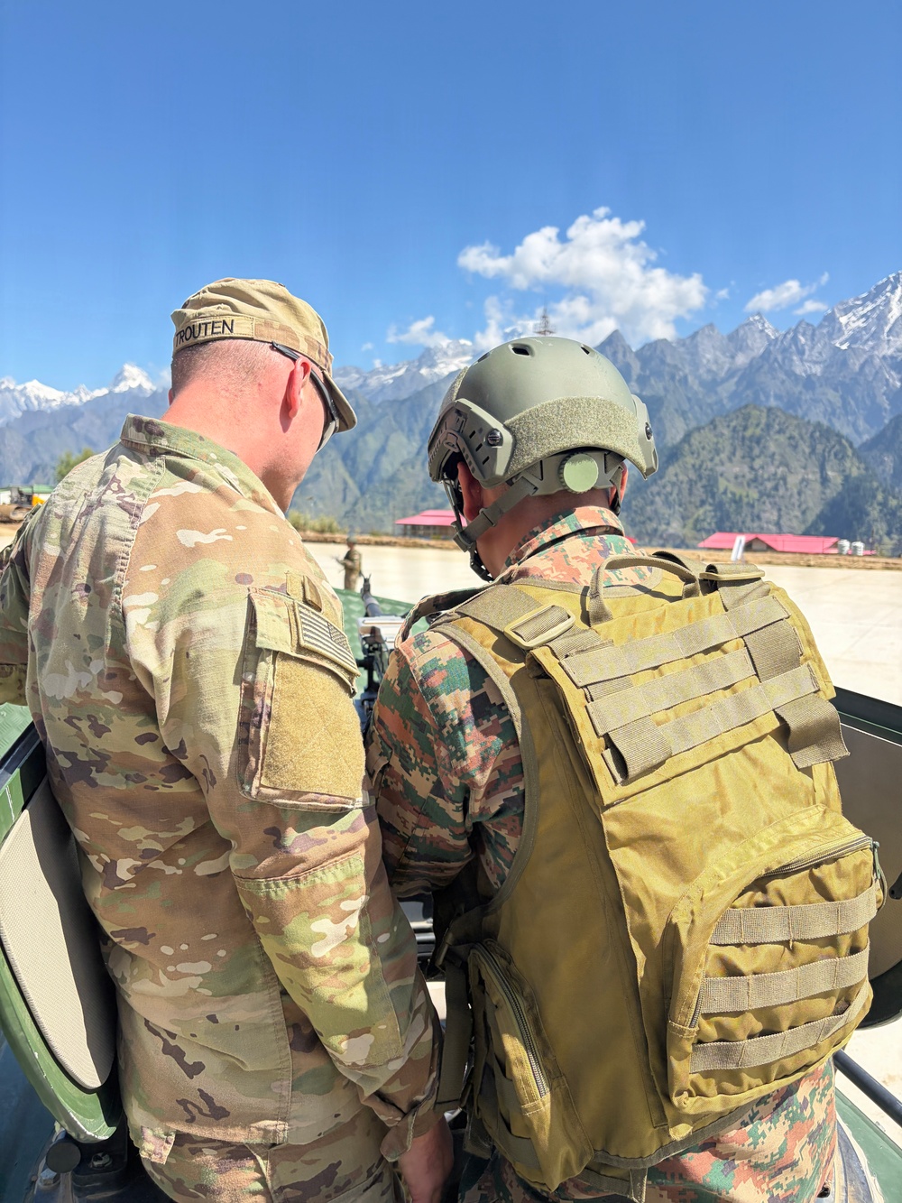 Two military personnel are photographed from behind in a mountainous setting with snow-capped peaks visible in the background under a clear blue sky. One soldier wears US Army MultiCam (OCP) uniform with a name tape reading 'TROUTEN' and a US flag patch, while the second wears a different camouflage pattern with a tactical helmet and tan/coyote backpack. They appear to be engaged in a joint military activity, possibly a bilateral or multinational exercise.