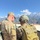 Yudh Abhyas 2026: U.S., Indian Soldiers exchange weapons, equipment knowledge in Auli Two military personnel are photographed from behind in a mountainous setting with snow-capped peaks visible in the background under a clear blue sky. One soldier wears US Army MultiCam (OCP) uniform with a name tape reading 'TROUTEN' and a US flag patch, while the second wears a different camouflage pattern with a tactical helmet and tan/coyote backpack. They appear to be engaged in a joint military activity, possibly a bilateral or multinational exercise.