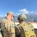 Two military personnel are photographed from behind in a mountainous setting with snow-capped peaks visible in the background under a clear blue sky. One soldier wears US Army MultiCam (OCP) uniform with a name tape reading 'TROUTEN' and a US flag patch, while the second wears a different camouflage pattern with a tactical helmet and tan/coyote backpack. They appear to be engaged in a joint military activity, possibly a bilateral or multinational exercise.