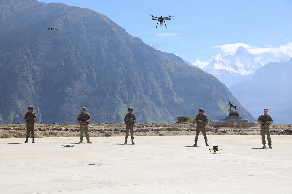 Five military personnel in camouflage uniforms are conducting a drone operation exercise on a concrete pad in a mountainous region. Multiple drones are visible — two airborne and at least two on the ground — suggesting a coordinated multi-drone training session. A monument or installation marker is visible in the background, and snow-capped peaks are visible in the distance.