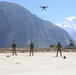 Five military personnel in camouflage uniforms are conducting a drone operation exercise on a concrete pad in a mountainous region. Multiple drones are visible — two airborne and at least two on the ground — suggesting a coordinated multi-drone training session. A monument or installation marker is visible in the background, and snow-capped peaks are visible in the distance.