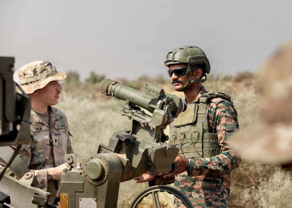 Two military personnel are interacting beside a crew-served weapon system, likely a TOW missile launcher or similar anti-tank guided missile (ATGM) system, in an arid outdoor training environment. One soldier wears U.S. Army OCP (Operational Camouflage Pattern) uniform with a patrol cap, while the other wears a different camouflage pattern with a tactical helmet and body armor, suggesting a joint or combined arms exercise. The setting and uniforms indicate a multinational military training or advisory scenario.