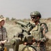 Two military personnel are interacting beside a crew-served weapon system, likely a TOW missile launcher or similar anti-tank guided missile (ATGM) system, in an arid outdoor training environment. One soldier wears U.S. Army OCP (Operational Camouflage Pattern) uniform with a patrol cap, while the other wears a different camouflage pattern with a tactical helmet and body armor, suggesting a joint or combined arms exercise. The setting and uniforms indicate a multinational military training or advisory scenario.