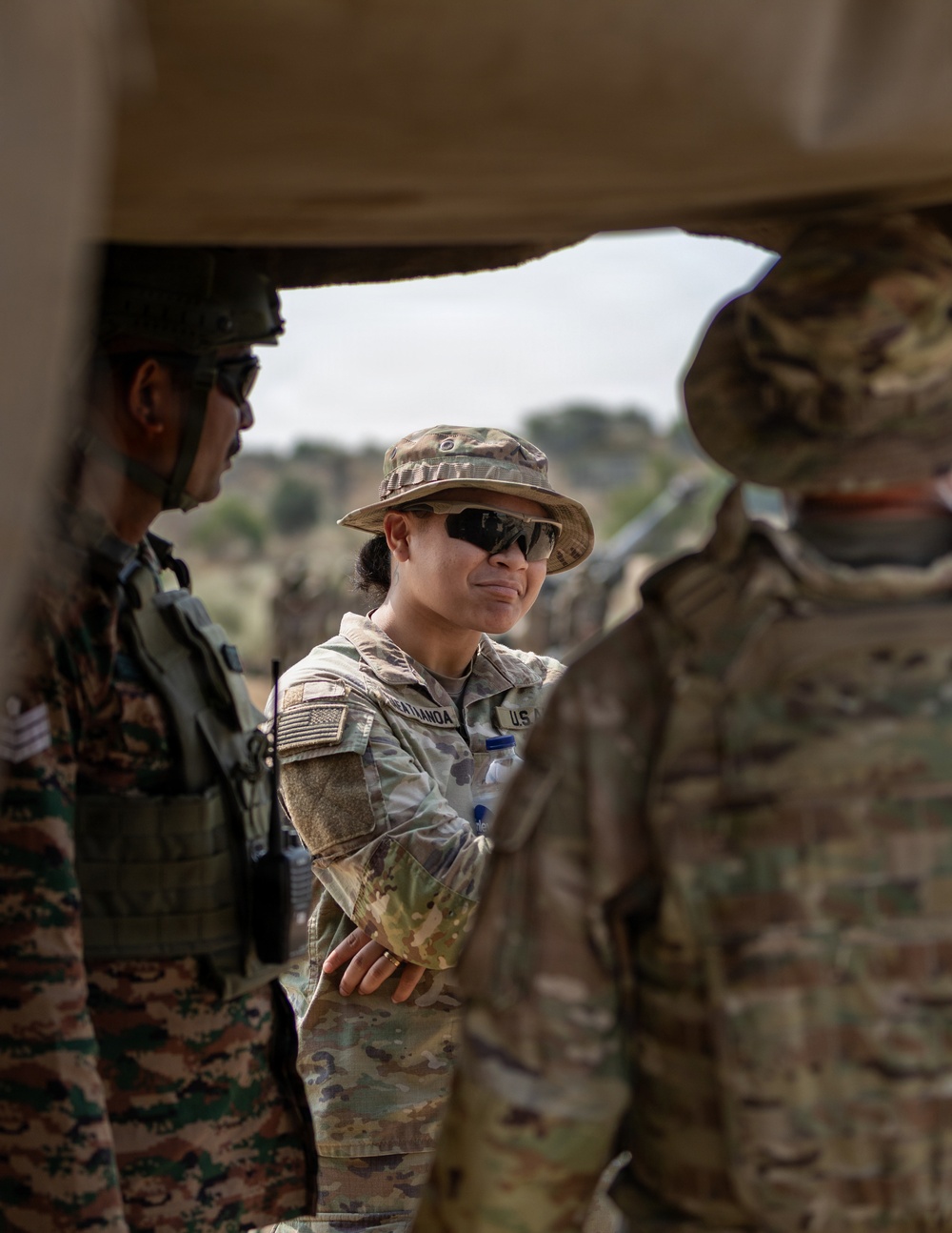 The image shows military personnel from at least two different armed forces engaged in what appears to be a briefing or discussion in an outdoor field environment. A central figure wearing U.S. Army Operational Camouflage Pattern (OCP) uniform with a boonie hat, tactical sunglasses, and a visible U.S. flag patch is interacting with personnel in a different camouflage pattern. The scene is framed through a vehicle or structure opening, suggesting a field exercise or joint military operation.