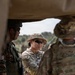 The image shows military personnel from at least two different armed forces engaged in what appears to be a briefing or discussion in an outdoor field environment. A central figure wearing U.S. Army Operational Camouflage Pattern (OCP) uniform with a boonie hat, tactical sunglasses, and a visible U.S. flag patch is interacting with personnel in a different camouflage pattern. The scene is framed through a vehicle or structure opening, suggesting a field exercise or joint military operation.