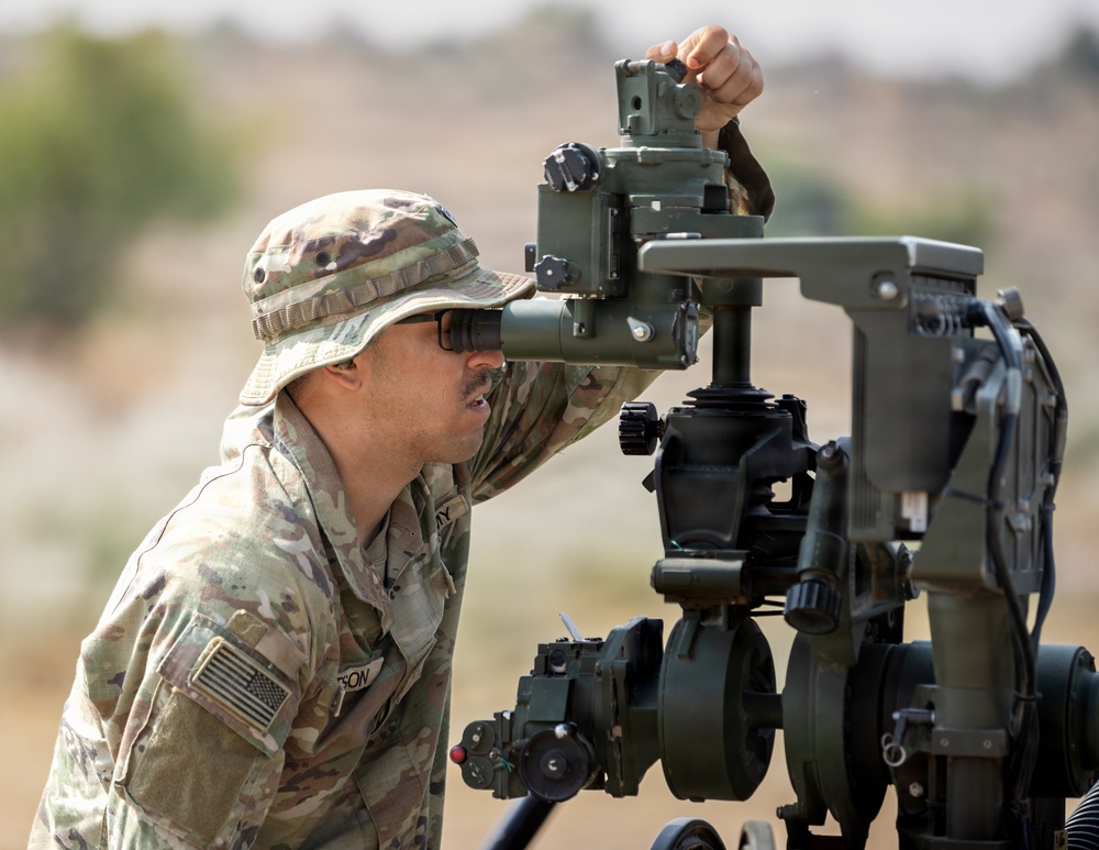 A U.S. Army soldier in MultiCam/OCP camouflage uniform and boonie hat is looking through the sight of an artillery piece, likely an M777 howitzer or similar field artillery system. The soldier is actively adjusting or aiming the weapon system using the panoramic telescope/sight mounted on the breech assembly. The setting appears to be an arid, open training range environment.