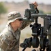 A U.S. Army soldier in MultiCam/OCP camouflage uniform and boonie hat is looking through the sight of an artillery piece, likely an M777 howitzer or similar field artillery system. The soldier is actively adjusting or aiming the weapon system using the panoramic telescope/sight mounted on the breech assembly. The setting appears to be an arid, open training range environment.
