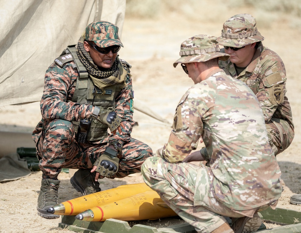 Three military personnel are gathered around a yellow artillery round or munition on the ground in an outdoor field environment. Two soldiers wearing U.S. Army MultiCam (OCP) uniforms are interacting with a third soldier in a different camouflage pattern, likely from a partner nation's military. The scene appears to depict a joint training or familiarization exercise involving artillery ammunition.