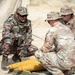 Three military personnel are gathered around a yellow artillery round or munition on the ground in an outdoor field environment. Two soldiers wearing U.S. Army MultiCam (OCP) uniforms are interacting with a third soldier in a different camouflage pattern, likely from a partner nation's military. The scene appears to depict a joint training or familiarization exercise involving artillery ammunition.