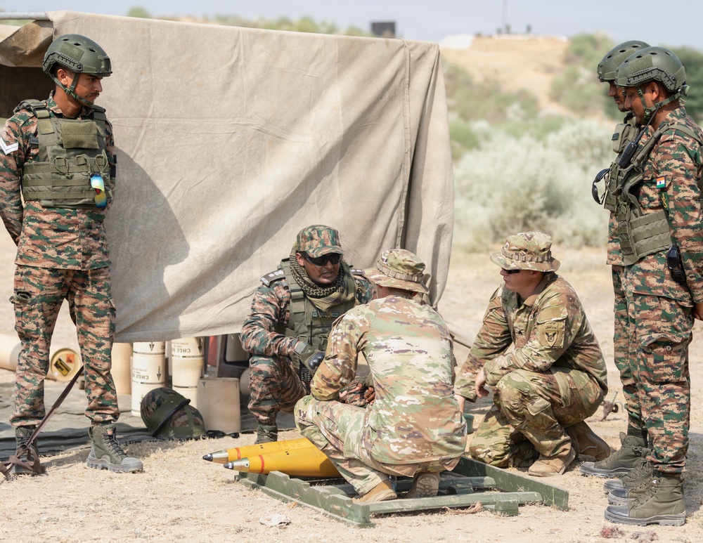 A group of military personnel from at least two different nations are gathered around a yellow munition/ordnance item placed on a wooden pallet on arid ground. Soldiers wearing distinctive pixelated camouflage uniforms with what appears to be an Indian flag patch are interacting with soldiers in OCP (Operational Camouflage Pattern) uniforms, suggesting a joint military exercise or training event. The setting appears to be an outdoor training area with a canvas shelter in the background.