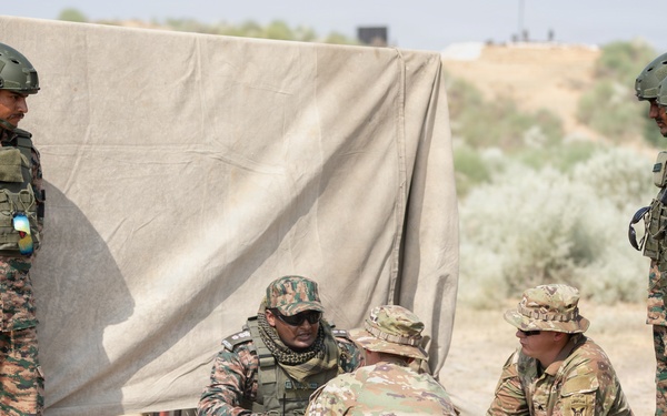 A group of military personnel from at least two different nations are gathered around a yellow munition/ordnance item placed on a wooden pallet on arid ground. Soldiers wearing distinctive pixelated camouflage uniforms with what appears to be an Indian flag patch are interacting with soldiers in OCP (Operational Camouflage Pattern) uniforms, suggesting a joint military exercise or training event. The setting appears to be an outdoor training area with a canvas shelter in the background.