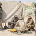 A group of military personnel from at least two different nations are gathered around a yellow munition/ordnance item placed on a wooden pallet on arid ground. Soldiers wearing distinctive pixelated camouflage uniforms with what appears to be an Indian flag patch are interacting with soldiers in OCP (Operational Camouflage Pattern) uniforms, suggesting a joint military exercise or training event. The setting appears to be an outdoor training area with a canvas shelter in the background.
