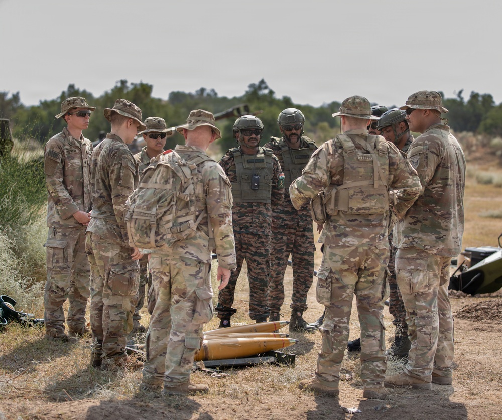 A group of military personnel in camouflage uniforms are gathered in an outdoor arid training environment, appearing to conduct a briefing or instruction session. Two individuals in the center wear distinctive darker camouflage uniforms with body armor and helmets, suggesting they may be from a different military force than the others. Artillery shells or large munitions are visible on the ground in the foreground.